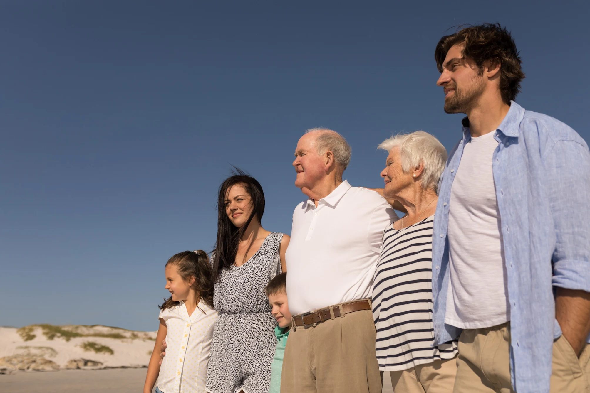 Low angle view of happy multi-generation family with arms around standing on beach in the sunshine